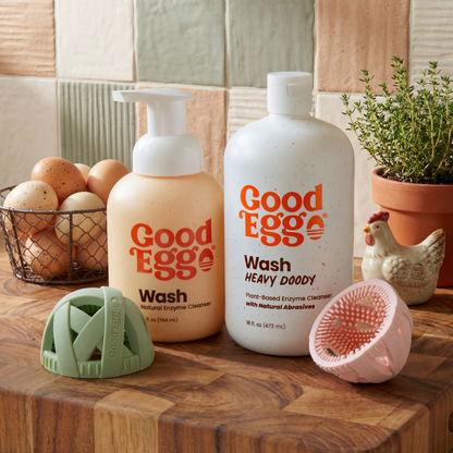 Two bottles of GoodEgg wash on a wooden surface with GoodEgg Brushes and kitchen items in the background.